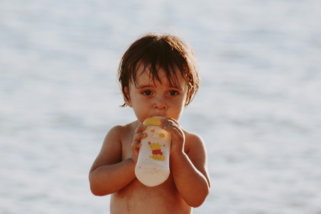 A cute child sipping from a bottle at the beach, showcasing innocence and playfulness.