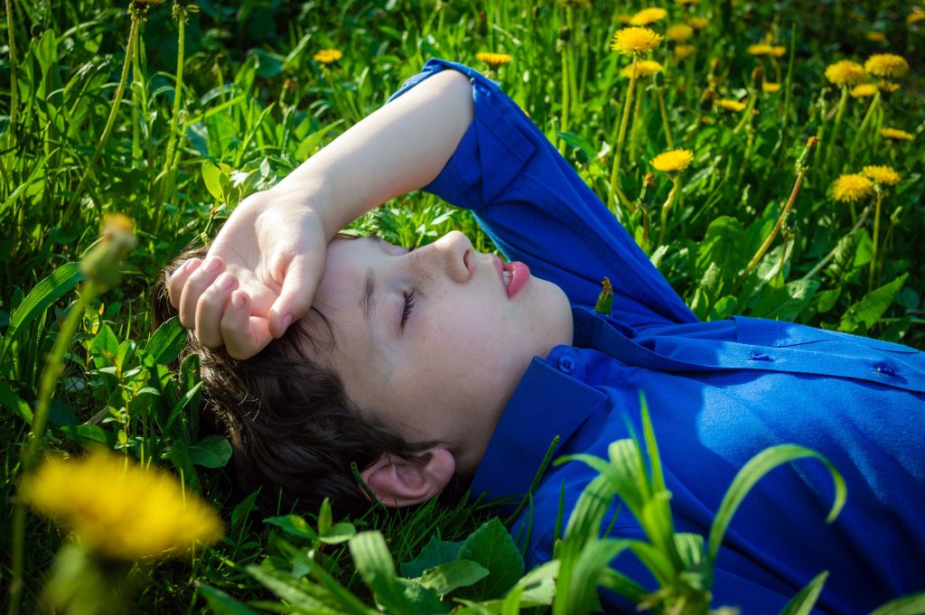 A young boy in a blue shirt lays in a lush field of dandelions with eyes closed, enjoying a peaceful moment.